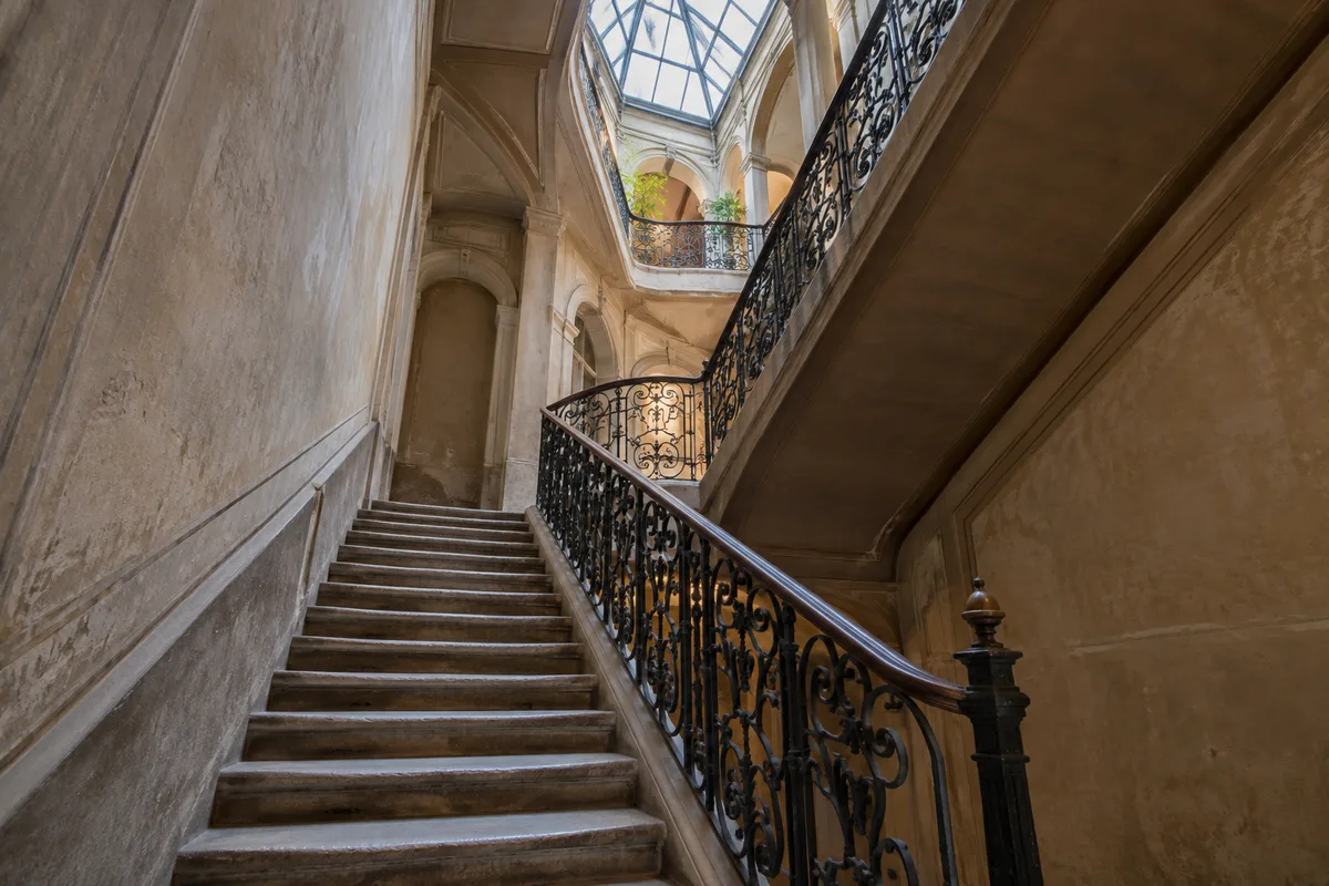 Cage d’escalier ancienne avec rampe en fer forgé et marches en pierre usées, lumière tombant d’une verrière